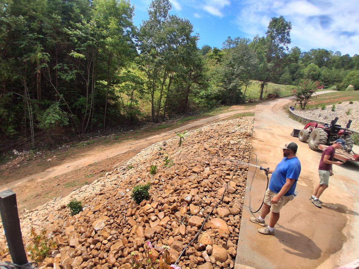 The crew watering in new plantings on a river rock bed along a concrete drive, with woods beyond