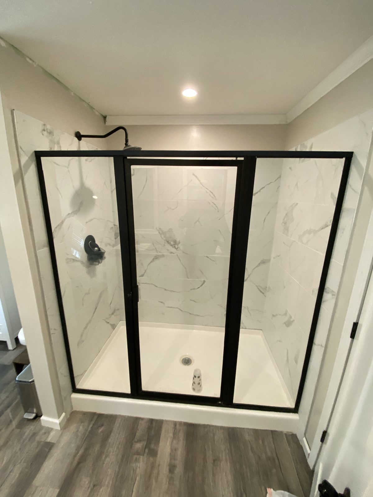 A new walk-in shower with white marble-look tile walls, a white shower base and a black-framed glass door, above gray plank flooring
